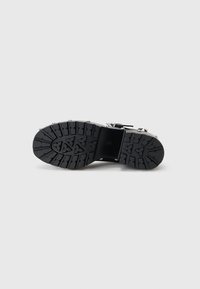 Black rubber sole with textured tread pattern and metal studs. Flat design with a rounded toe. Smooth leather upper with adjustable strap.