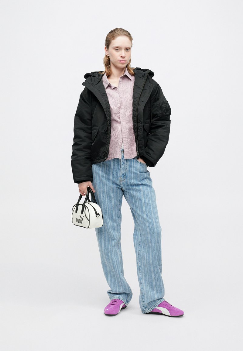 Schwarze Daunenjacke, hellrosa Hemd mit Knöpfen, blau gestreifte Jeans, lila Schuhe und eine kleine weiße Handtasche mit schwarzen Akzenten.