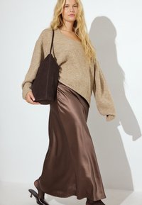 Beige Wollsweater mit Ballonärmeln, kombiniert mit einem braunen Satin-Maxikleid. Das Model hält eine dunkelbraune, strukturierte Tasche und trägt schwarze High Heels.