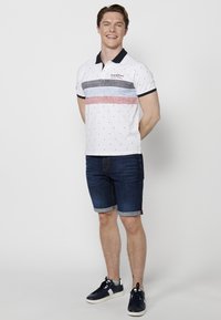 Bela polo majica kratkih rukava s horizontalnimi modrimi in rdečimi črtami, kombinirana s temnimi denim kratkimi hlačami in mornarsko modrimi supergami. Preprost geometrijski vzorec.
