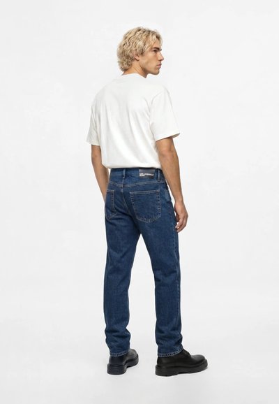Homme blond portant un t-shirt blanc uni, un jean bleu foncé et des chaussures noires, debout et regardant sur le côté devant un fond blanc uni.