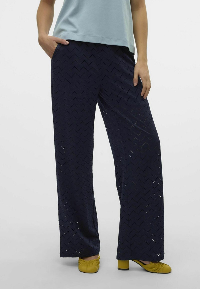 Navy siniset leveälahkeiset housut, joissa on chevron-kuviointi ja leikkuureunat. Yhdistetty keltaisiin jalkineisiin neutraalilla taustalla.