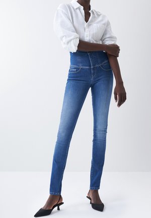 Person wearing high-waist blue skinny jeans, white button-up shirt with rolled sleeves, and black low-heeled slingback shoes standing.
