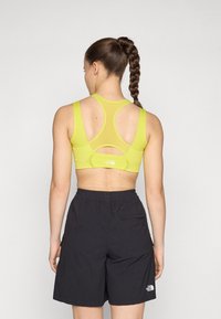 Sujetador deportivo amarillo con un respaldo de malla y detalle recortado, combinado con pantalones cortos negros que tienen una cinturilla elástica y un bolsillo lateral.