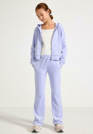 Hellviolette Kapuzenjacke zum Reißverschluss mit zwei Fronttaschen, kombiniert mit passenden Jogginghosen. Glatter Stoff mit Kordelzug an der Taille. Sneakers mit Polsterung.