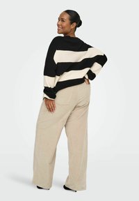 Pletena majica s crnim i kremastim prugama, dugih rukava, uparena s bež manšestarskim hlačama širokih nogavica. Model stoji bočno, ruke na bokovima.