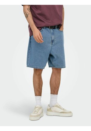 Homme portant un short en jean bleu ample, des baskets blanches, des chaussettes blanches et un t-shirt bordeaux, debout les mains partiellement dans les poches.