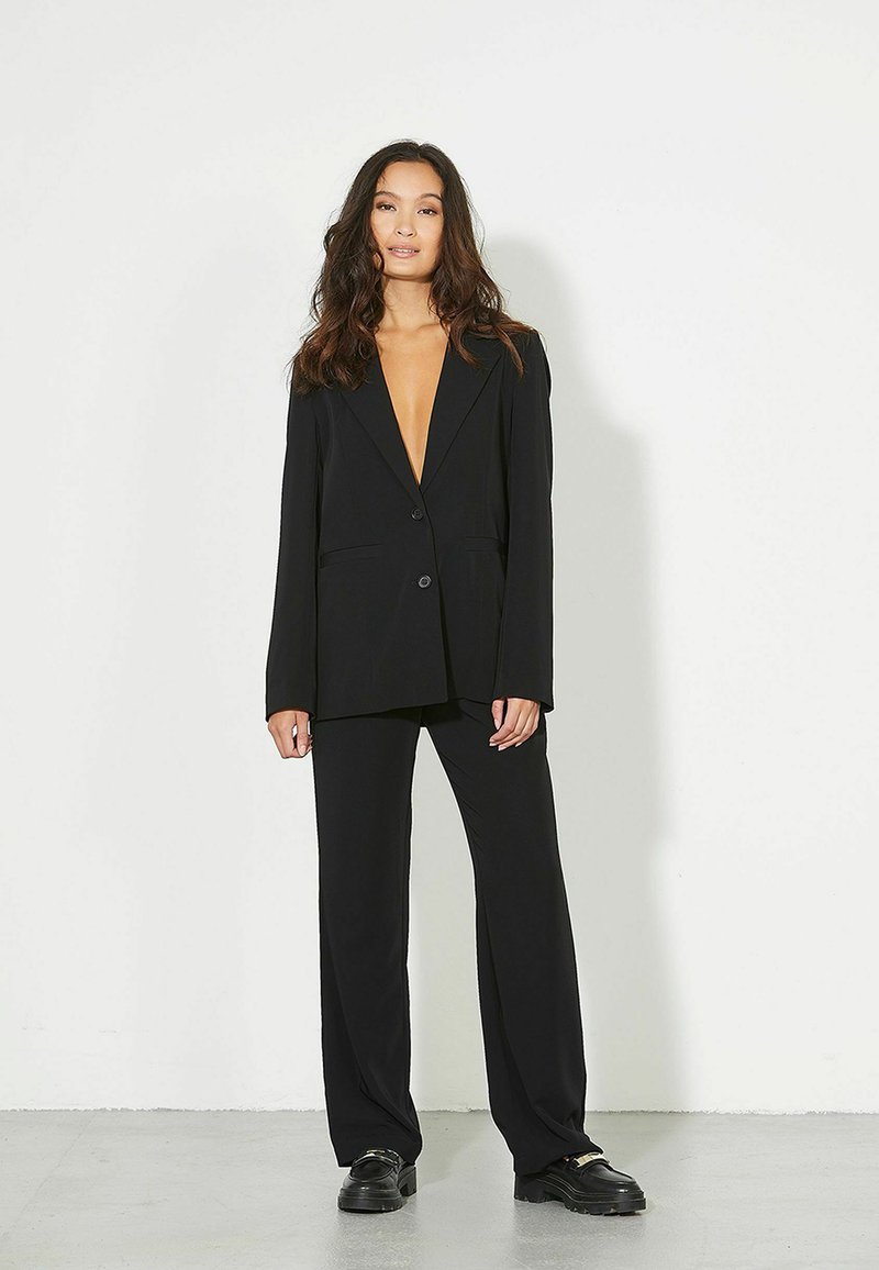 Γυναίκα με μακριά κυματιστά μαλλιά, ντυμένη με μαύρο φαρδύ σακάκι και αντίστοιχο παντελόνι με φαρδιά πόδια, στέκεται μπροστά σε έναν λευκό απλό τοίχο.