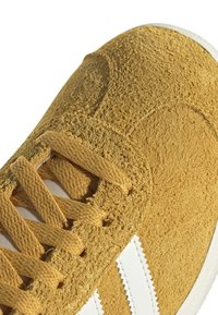 Žlutá semišová teniska s bílými koženými pruhy, strukturovaným svrškem a plochými tkaničkami. Zaoblená špička s bílou gumovou podrážkou.