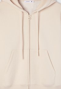 Beżowa bluza z kapturem na zamek, charakteryzująca się miękką fakturą, kieszeniami z przodu, sznurkami reguluącymi oraz ribbowanym dołem. Metka przedstawia nazwę marki "Terranova" oraz rozmiar "S."