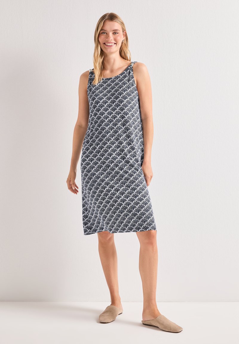 Femme souriante debout portant une robe sans manches noire et blanche à motifs jusqu'aux genoux et des chaussures beiges à enfiler sur un fond uni.