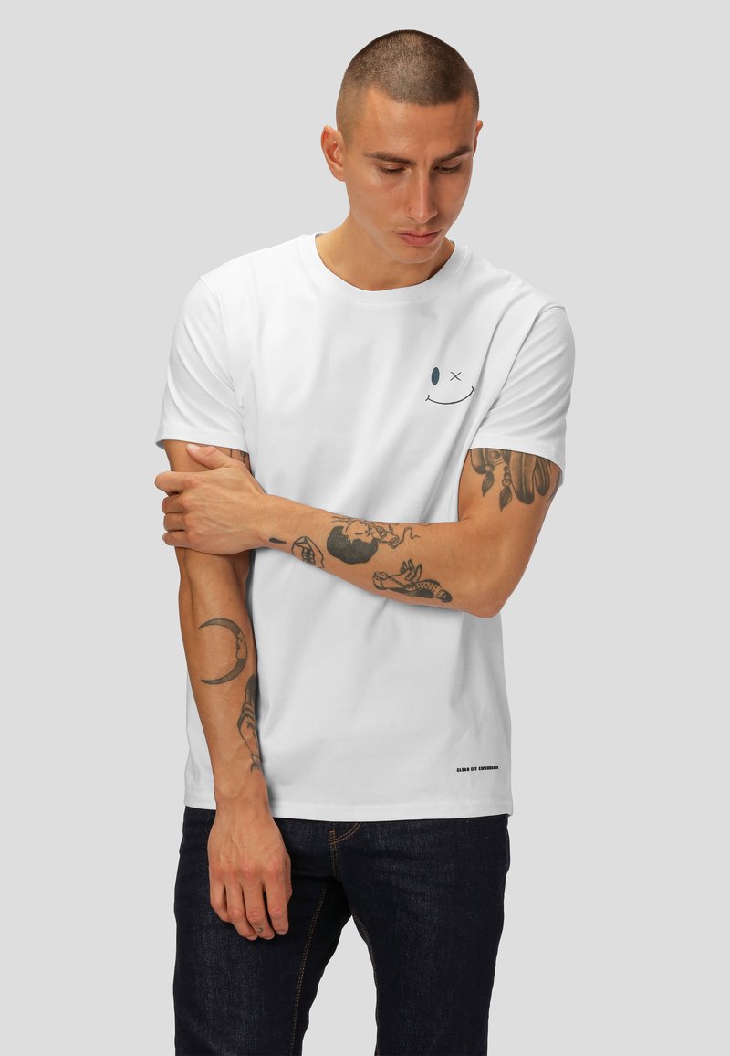 T-shirt en coton blanc à manches courtes, avec un motif de visage souriant bleu sur la poitrine et une étiquette noire en bas.