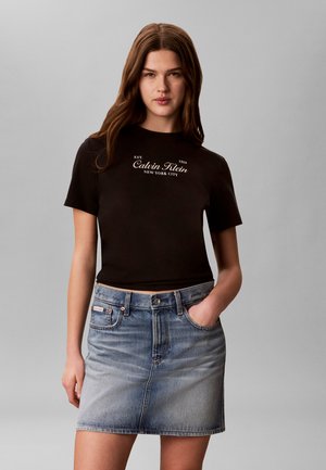 Jeune femme portant un t-shirt noir avec le logo Calvin Klein et une jupe en jean bleue, debout avec une main dans la poche devant un fond gris.