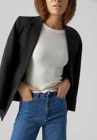 Kvinna i svart blazer, vit ribbad ärmlös topp och blå jeans med händerna i fickorna mot en enfärgad bakgrund.