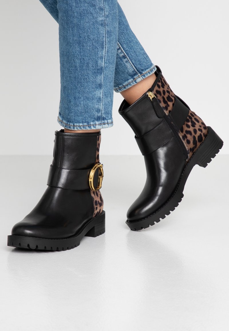 Bottes noires chevilles avec boucle dorée et accents imprimé léopard portées avec un jean bleu sur un sol blanc.