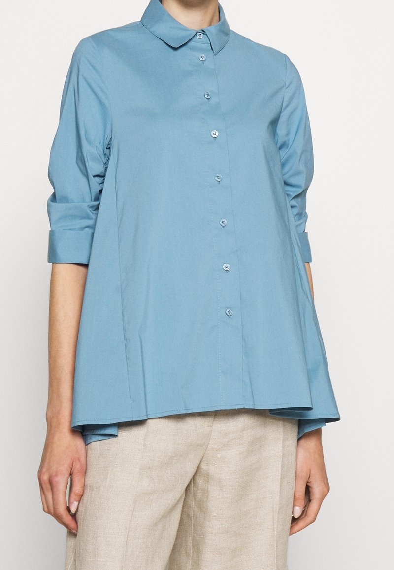 Personne portant une chemise bleu clair ample à boutons avec des manches trois-quarts et un pantalon beige, montrée des épaules aux hanches.