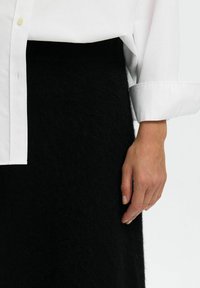 Balta krekla blūze, kas valkāta pār melnu, teksturētu vilnas svārku, ar redzamu roku, kas atpūšas tuvu svārka malai.