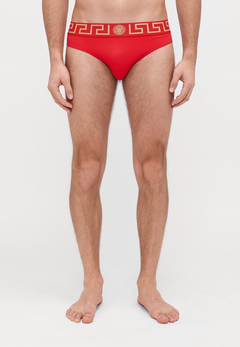 Rote Herren-Schwimmslips mit einem goldenen griechischen Schlüssel-Muster am Bund und einem zentralen goldenen Logo-Akzent, aus glattem, dehnbarem Stoff.