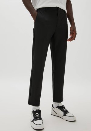 Homme portant un pantalon noir droit, des chaussettes blanches et des baskets blanches avec des lacets noirs, debout avec une main dans la poche.
