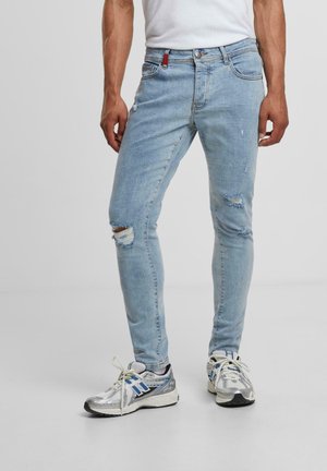 Lyseblå denim jeans med slidte detaljer, slim fit, og en rød mærkatdetalje nær taljen. Sat sammen med sølvfarvede sneakers.