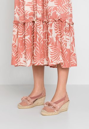Ben iført ferskenfarvet midi-nederdel med blomstermønster og lyserøde kileespadriller med stofblomster og elastiske stropper på beige gulv.