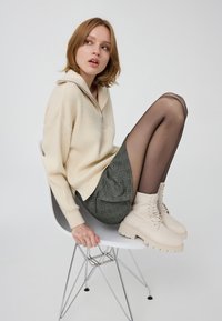 Kremowy prążkowany sweter z kołnierzem na zamek, zielona kraciasta spódnica, przezroczyste czarne rajstopy i beżowe buty sznurowane, siedząca na białym krześle.