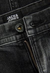Mustad teksased, millel on nähtav lukku, brändi silt "JACK & JONES" ja metallist nööpkinnitus. Sile tekstuur koos õmbluste detailidega.