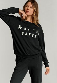 Čierna mikina s vaflovou pleteninou a voľným strihom. Na prednej strane je nápis "b by TED BAKER" v bielej farbe. Môže sa kombinovať s čiernymi nohavicami.