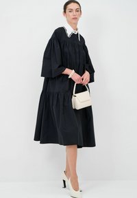 Zwarte, gelaagde, lange mouwen jurk met een witte kantkraag; gecombineerd met een witte handtas en beige hoge hakken.