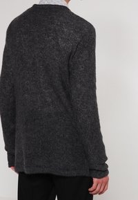 Pull en maille grise avec une coupe ample, tissu texturé, épaules tombantes et dos légèrement allongé. Design décontracté avec manches longues.