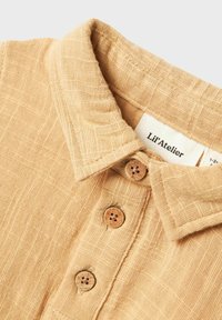 Camicia di lino beige con colletto, dotata di cinque bottoni in legno lungo il patta e un motivo superficiale testurizzato. Etichetta visibile.