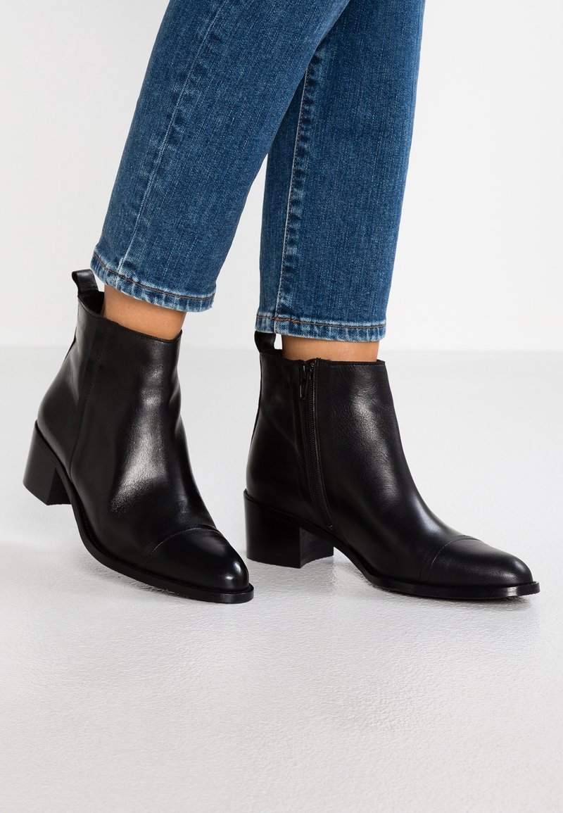Stivaletti neri in pelle con punta affusolata e zip laterale, caratterizzati da un tacco a blocco e una texture liscia. Abbinati a jeans blu a vita bassa.