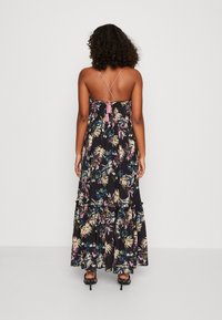 Maxikleid mit schwarzem Hintergrund, ausgestattet mit einem mehrfarbigen tropischen Blattdruck, gestuften Rüschen und über Kreuz verlaufenden Spaghettiträgern mit einer Quasten-Verzierung.