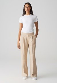 Witte, aansluitende T-shirt in combinatie met lichtbeige, op maat gemaakte broeken met plooien, zijzakken en witte sneakers. Soepele, strakke uitstraling.