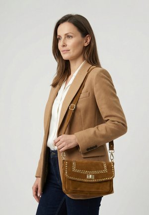 Woman wearing camel blazer and white blouse, carrying brown studded suede shoulder bag with adjustable strap.
