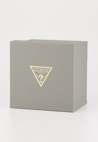 Boîte grise texturée avec un logo triangulaire doré affichant "GUESS" et un point d'interrogation. Fermée, avec des coins vifs et sans motifs visibles.