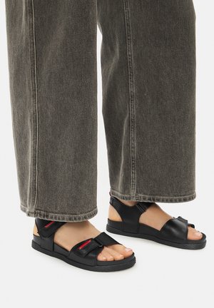 Čierne sandále s nastaviteľnými popruhmi, textúrovanou podrážkou a výrazným prstovým pásikom. Nosia sa s šedými širokými džínsami, odhaľujúcimi bosé nohy.