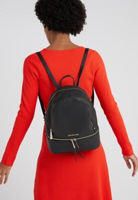 Sac à dos en cuir noir avec des détails en or, de forme arrondie, doté de doubles zips et d'une poignée sur le dessus. Porté sur une robe rouge à côtes.