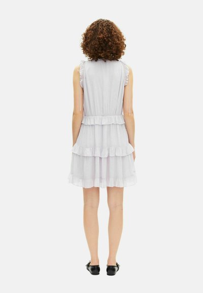 Vrouw met krullend haar staat met haar rug naar de camera, draagt een mouwloze witte jurk met gelaagde richelletjes en zwarte platte schoenen op een witte achtergrond.