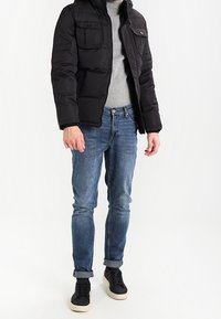 Svart puffjacka med framfickor, grå tröja, blå denimjeans och svarta sneakers med vita sulor. Avslappnat outfit.