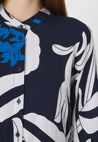 Camisa azul-marinho com botões, apresentando grandes padrões florais brancos e azuis, feita de um tecido suave com ligeiro brilho e gola arredondada.