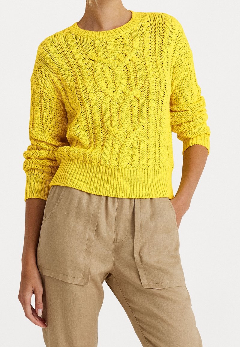Femme portant un pull jaune vif à torsades avec poignets et ourlet côtelés, associé à un pantalon beige taille haute avec poches avant.