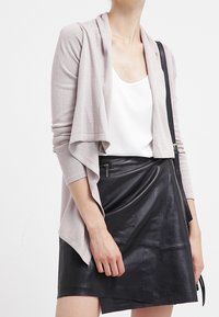 Lichtgrijze cardigan met een gedrapeerde voorkant, gedragen over een witte top en een zwarte neplederen rok met een diagonale snit en ritsdetails.
