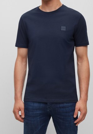 Mężczyzna noszący prosty granatowy t-shirt z krótkim rękawem z małym wytłoczonym logo na lewej piersi, w połączeniu z ciemnoniebieskimi dżinsami.