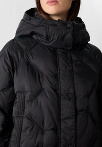 Kobieta ubrana w czarną pikowaną kurtkę puchową z wysokim kołnierzem i zatrzaskami, pokazująca jedynie górną część torsu i usta.