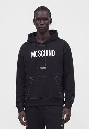 Ung mand iført sort Moschino hoodie med hvid tekst "MOSCHINO Milano" og matchende sorte bukser mod en ensfarvet grå baggrund.