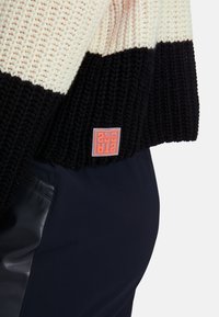 Strickpullover mit cremefarbenen, schwarzen und pfirsichfarbenen Streifen; gerippte Textur; kleines oranges Logo-Patch; kombiniert mit navyblauen Hosen.