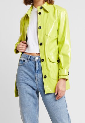 Person wearing shiny lime green buttoned coat over white cropped top and light blue high-waist jeans, standing against plain background.