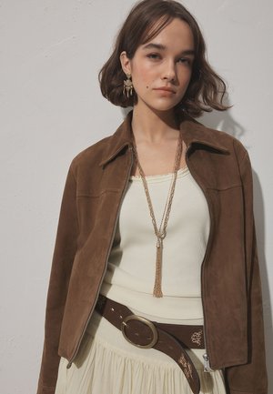 Junge Frau mit kurzen braunen Haaren, die eine braune Wildlederjacke, ein beiges Top, einen gemusterten braunen Gürtel, eine lange Halskette und hängende Ohrringe trägt.