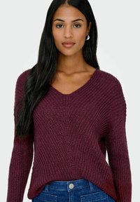 Maglione borgogna lavorato a maglia con scollo a V, texture a coste, spalle scese e orlo leggermente curvo. Abbinato a jeans blu scuro.
